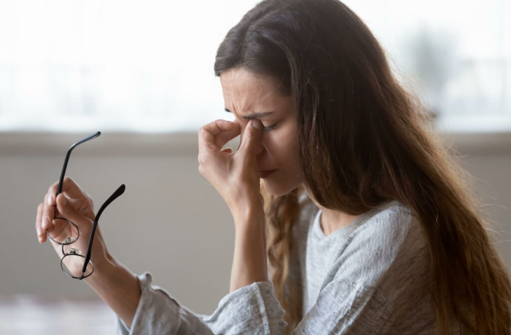 A young adult taking off her glasses to rub her eyes due to discomfort from her meibomian gland dysfunction.