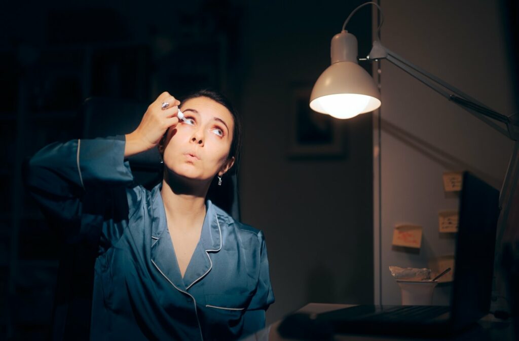 A young adult in pyjamas applies eyedrops to red eyes to relieve dry eyes while working in a dim room at night