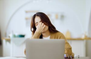 An adult takes a break from working on their computer and pinches the bridge of their nose as they feel a tension headache from eye strain coming on.