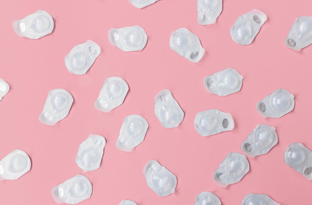 Daily disposable contact lenses in blister packaging are strewn across a bright pink background.