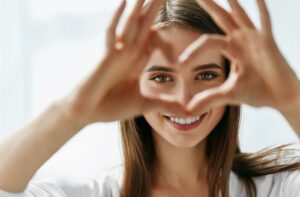 A person smiling and framing their eye with their hands in a heart shape, illustrating healthy vision and eye comfort.