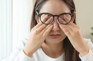 A person wearing black-rimmed glasses and using their fingers to rub their closed eyelids, showing dry eye fatigue.