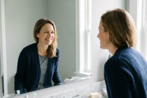 Person in a navy cardigan smiling at their reflection in a bathroom mirror.