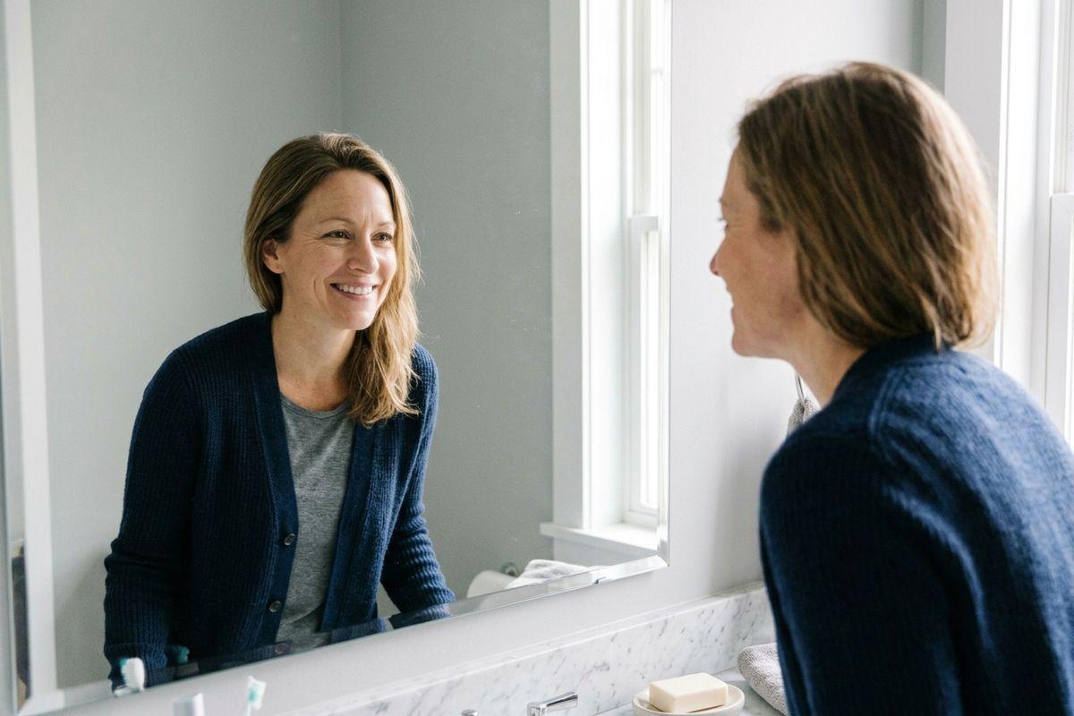 Person in a navy cardigan smiling at their reflection in a bathroom mirror.
